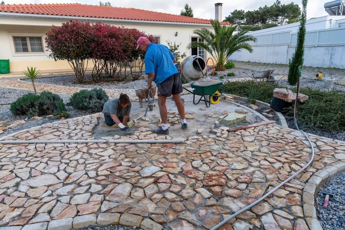 Michael Straub laying a natural crazy-paving stone patio with a cement mixer and wheelbarrow at a Silver Coast property, with a younger team member placing stones